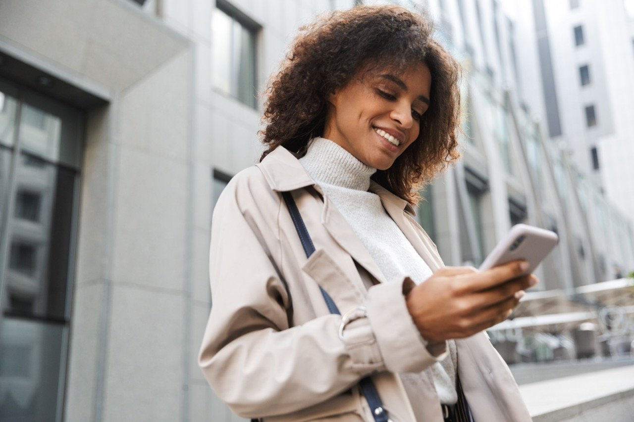african american woman with curly hair in white coat and sweater smiling look at phone