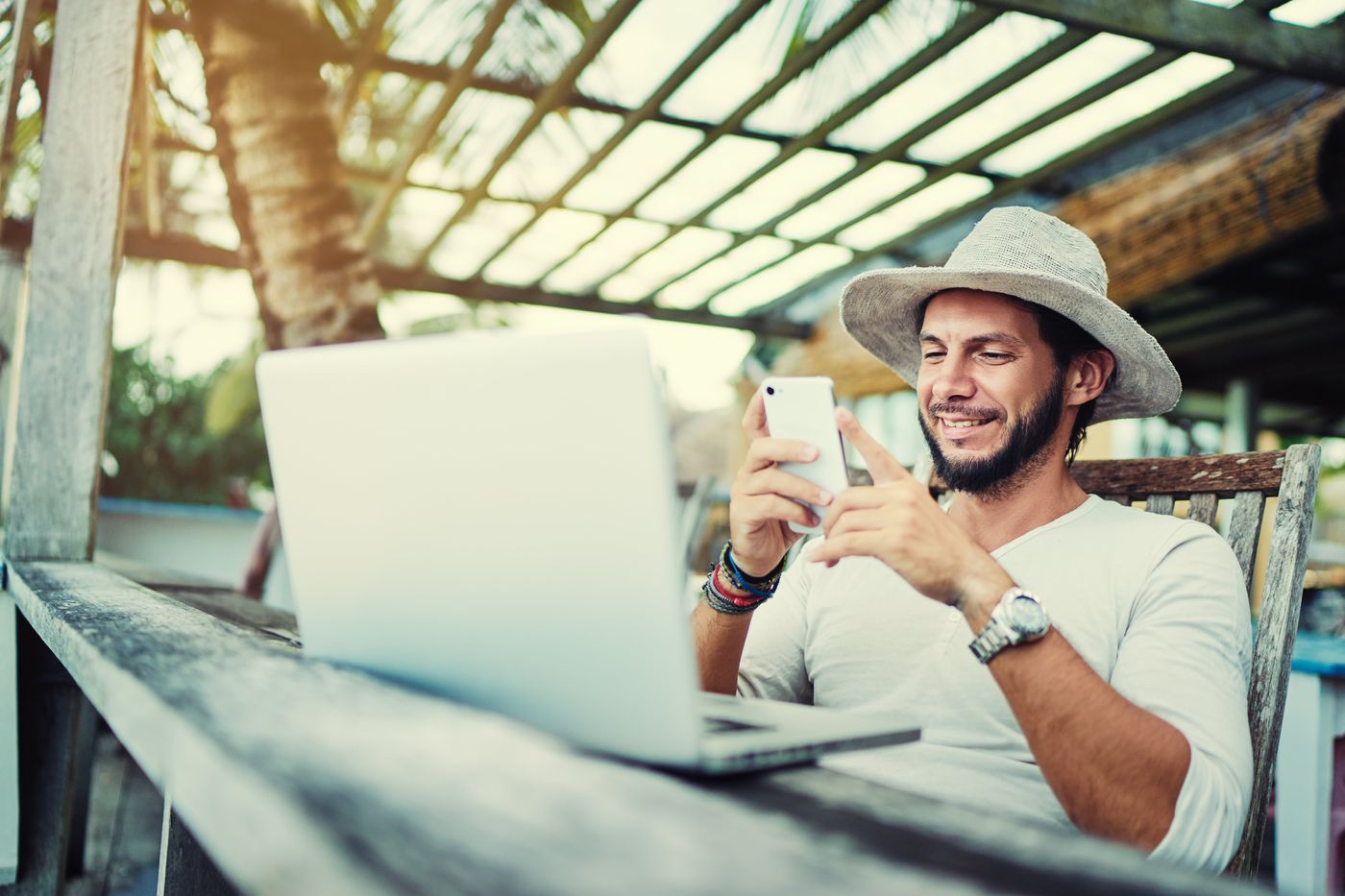 middle aged man in white hat on vacation smiling looking at phone with laptop in front of him