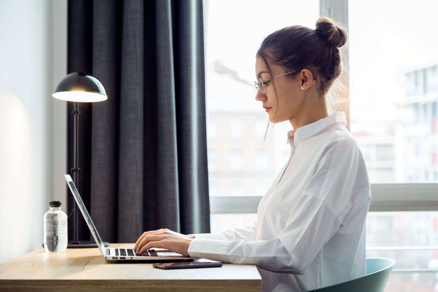 side shot of woman in white button up working at laptop in hotel room